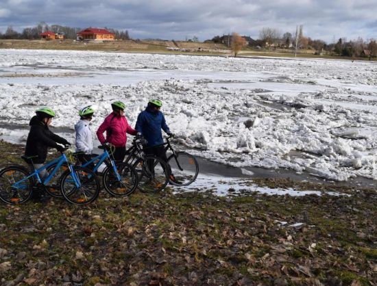 Četri riteņbraucēji ar ķiverēm un spilgtās jakās apstāju&scaron;ies Rundāles ūdensdzirnavu dīķa krastā. Skats uz pla&scaron;u, ledus krāvumiem klātu ūdens virsmu pavasara palu laikā. Fonā redzamas Rundāles atpūtas centra komplekss. 