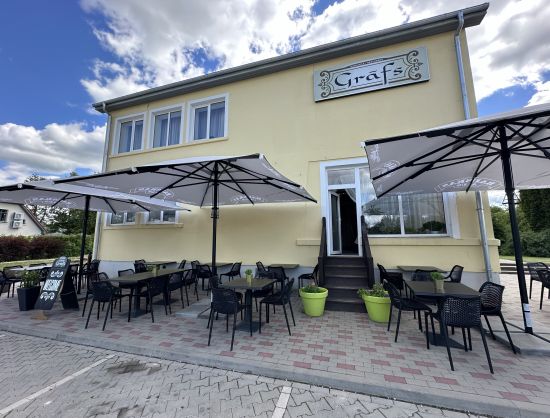 Kafejnīcas 'Grāfs' āra terase vasaras sezonā: galdiņi ar saulessargiem un ērtiem krēsliem, ko ieskauj Rundāles pils vēsturiskā vide un sakoptā apkārtne.