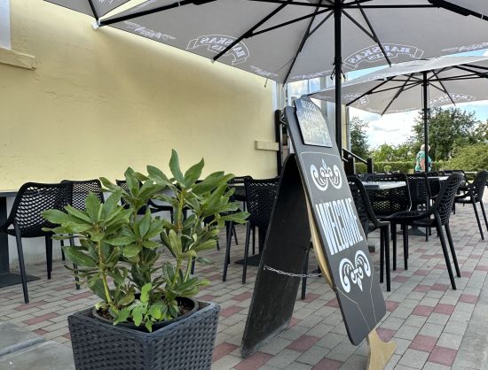 Kafejnīcas 'Grāfs' āra terase vasaras sezonā: galdiņi ar saulessargiem, redzams dekoratīvs sagaidī&scaron;anas baneris 'Welcome' pie ieejas