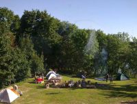 Rundāles atpūtas centrs/ Rundāles kempings vasaras dienā. Zaļā zālājā izvietotas vairākas dažādu krāsu teltis, viesi bauda atpūtu. Redzama iekārtota ugunskura vieta ar dūmiem un āra mēbelēm, ko ieskauj kupli lapu koki. Fonā pavīd ūdens virsma