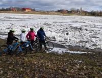 Četri riteņbraucēji ar ķiverēm un spilgtās jakās apstāju&scaron;ies Rundāles ūdensdzirnavu dīķa krastā. Skats uz pla&scaron;u, ledus krāvumiem klātu ūdens virsmu pavasara palu laikā. Fonā redzamas Rundāles atpūtas centra komplekss. 